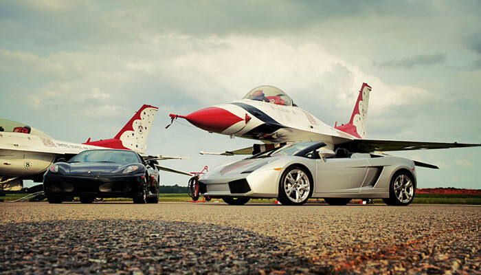 fway-2025-precision-exotics Precision Exotics - Two exotics sports cars, one black, one silver, parked on the tarmac, in front of a two white, red, and blue fighter jets
